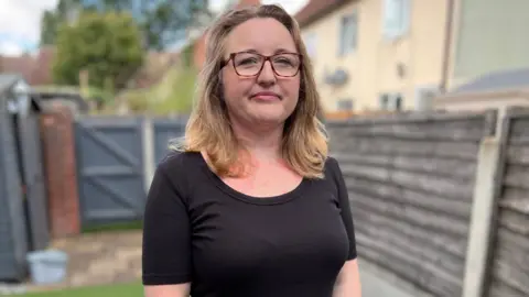 Stuart Woodward/BBC A woman with blonde hair and glasses looks at the camera, while standing in a back garden with fence panels behind her out of focus. She wears a black top.