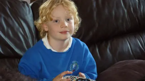Kai Madgwick Kai sits in a blue school jumper and white polo shirt holding a television remote and looking toward the camera 