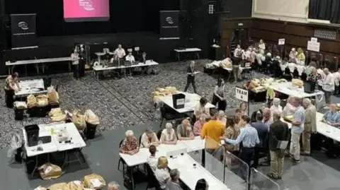 A bird's eye view of the Kent County Council election count in May 2025. Bags of ballots can be seen on tables around the hall, where a group of people are sat in a row counting the votes.