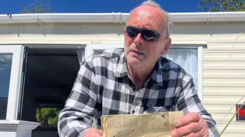 Walter Renwick is standing looking over a white fence surrounding a static caravan, holding a faded yellow newspaper. He is a man in his 60s wearning a black and white checked shirt and sunglasses. 