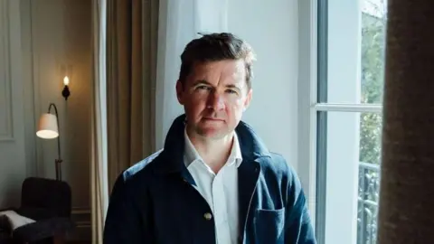 William Layzell pictured in a grand looking house, with wooden floors and a fireplace visible behind him. He's wearing a navy shacket with a white shirt, and has short brown hair.
