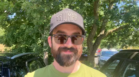 BBC Rich Collins is pictured wearing a dark grey cap with "Motorcycles Triumph" on the front. He is wearing sunglasses and has a brown beard. 