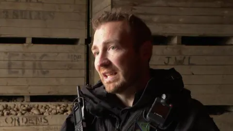 Sgt Rob Simpson, in his black police uniform with radio and bodycam equipment attached, speaks to the camera with crates of potatoes visible in the background. 