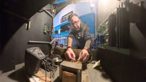 BBC/James Vincent An engineer kneels down behind an arcade machine with a multimeter in his hand. He's helping to check that Space Invaders is in full working order.