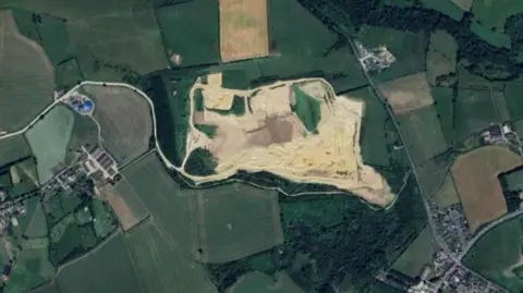 An aerial view of the quarry. It is a roughly rectangular light tan-coloured area surrounded by fields. A few buildings are to the left of the quarry and a large number of houses are to the right.