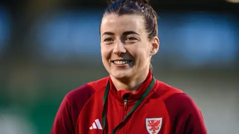 Getty Images Angharad James smiles at the camera. Her dark hair is tied back and she is wearing a red zip jacket with the Wales shield on one side and the Adidas logo on the other. 