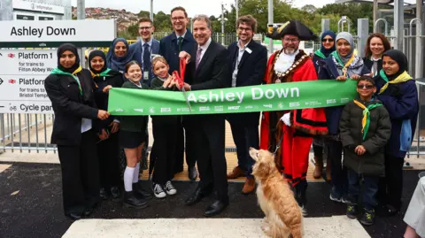 GWR Various dignitaries, including the mayor of bristol, the mayor of the west of england Dan Norris and a group of school children holding a pair of giant scissors in front of a banner with the words "Ashley Down", preparing to cut the banner