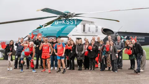 KSS Air Ambulance A group photo of Steve Wolfe and the KSS team. There are around 30 people in the photo all smiling at the camera. Behind the group is a KSS helicopter. 