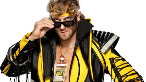 Getty Images Logan Paul with an elaborate yellow and black jacket, yellow headband and sunglasses which he is pulling down to show his eyes, and the Pokemon card in a jewelled clear case hanging around his neck on a chunky gold chain