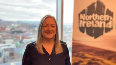 A blonde woman wearing a dark shirt smiles at the camera. Beside here is a sign which says Northern Ireland