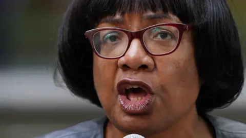 PA Media Labour MP Diane Abbott wears spectacles with maroon frames and speaks into a microphone
