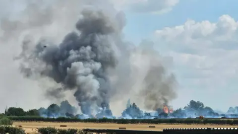Sharna Miller Large flames and plumes of smoke billow from brown fields lined with trees on a summer's day in the Wiltshire countryside