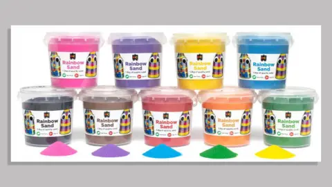 ACCC Nine different tubs of brightly coloured sand labelled as 'EC Rainbow Sand' are pictured against a white background. The colours are pink, purple, yellow, blue, black, chocolate brown, red, orange and dark green.