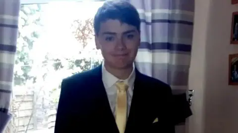 North Wales Police Ben Leonard smiling in front of a sunlit window wearing a black suit and yellow satin tie in a family photo