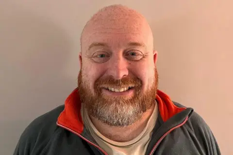 Jamie Lewis Jamie Lewis smiles at the camera showing his teeth. He is bald and has a ginger beard and blue eyes. He wears a grey sweatshirt and stands in front of a wall. 