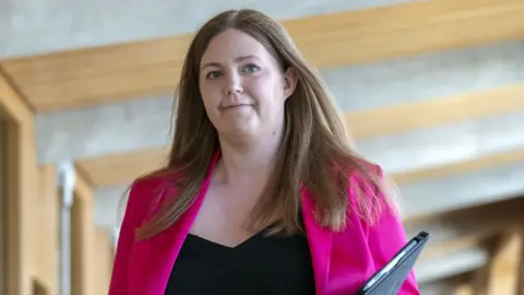 PA Media Gillian Mackay, with fair and wearing a pink jacket and black top, walks in the Scottish Parliament. She is visible from the chest up and is holding a black folder.