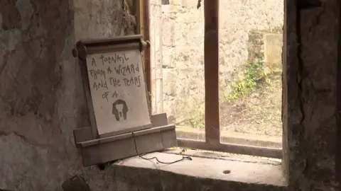 A scroll from the show leaning on a dusty windowsill in Gwyrch Caste. It says 'A toenail from a wizard and the tears of a ...'. Below the text is an image of an outline of a mummy's head.