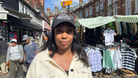 A woman in a cap in front of a market stall. She long black straight hair, and wears a cream coloured top. Behind her is a market stall selling men's shirts and polo shirts, and shoppers walking by.