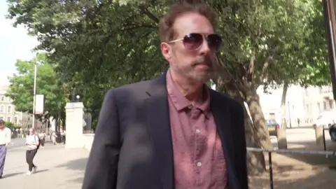 Chris Brain, a man wearing a salmon pink shirt, navy blue suit jacket and aviator sunglasses, walks on the pavement in the shade of trees outside Inner London Crown Court.