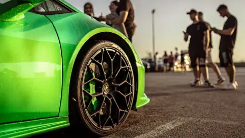 A stock image of a car meet, showing the front bumper of a bright green sports car in a car park, with attendees in the background blurred out.