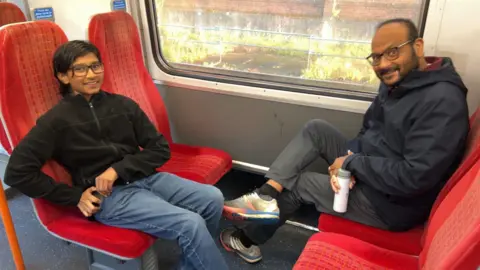 Father and son sitting on opposite train seats on a train. The father is holding a cup of a hot drink.