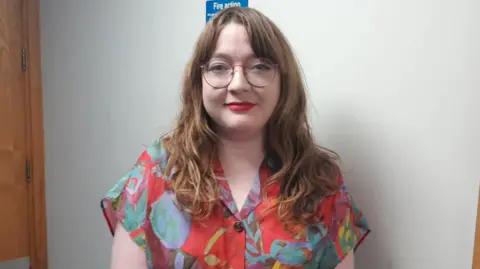 Áine Groogan - A woman with long hair wears a pair of red circular glasses and a red patterned shirt while standing in front of a white wall.