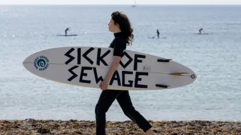 PA A woman carrying a surfboard that says 'Skk of sewage'