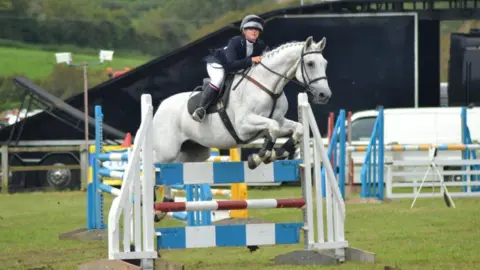 Ella Pallister is showjumping on a white horse. She is wearing a navy blue blazer with white leggings and knee high boots, with a black helmets. The horse has a braided mane and is jumping over a blue and white obstacle.