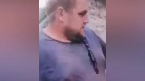 A picture of a man with a dark beard and a close-shaven head. He wears a dark blue t-shirt and has glasses hanging from the collar.