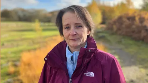 Dawn wearing a maroon North Face overgarment complete a ray bluish zip-up top, opinionated outdoors connected a grassy path. The inheritance features autumn foliage pinch aureate and orangish tones, scattered trees, and a somewhat cloudy sky.
