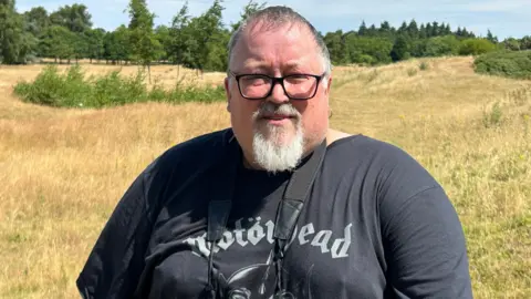 A man with short grey hair, a white goatee and black-rimmed glasses standing on a patch of grassland on a sunny day. Many trees are in the distance. He is wearing a black Motorhead t-shirt and has a pair of binoculars hanging around his neck.