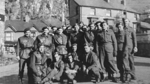 Black and white 1940s photo of group of soldiers standing in front of houses