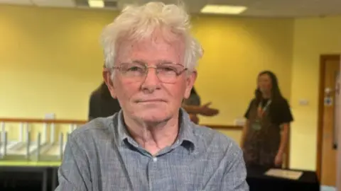 Molly Smith/BBC Ged Parker has grey hair and his wearing a grey shirt and glasses. There are people behind him out of focus.