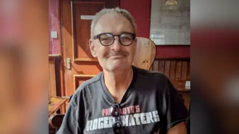 John Carr, a man with short receding grey hair, sits in a wood-panelled room. He wears a dark t-shirt with the words Pink Floyd's Roger Waters printed on it in capital letters, and black glasses. There is also a pair of black Ray-Ban sunglasses hanging from the neck of his t-shirt. He has a moustache and is smiling happily.