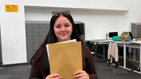 Lola smiles at the camera as she holds a manila envelop in front of her . she has brown hair and is a dark top