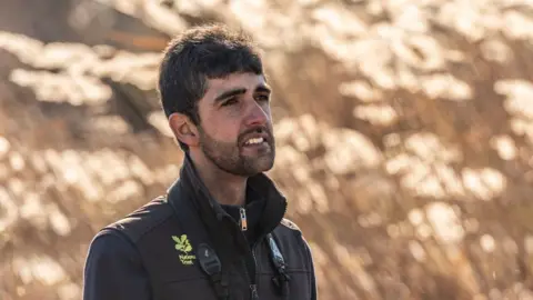 National Trust/Paul Morris Ajay Tegala standing somewhat side on and looking towards the right. He has dark hair and a close cropped black beard. He is wearing a black gilet, with a yellow National Trust logo, over a black top and is standing in front of blurred out reeds.