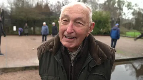 Ian Palmer/BBC Reginald Jones, wearing a brown and green jacket, stood in front of a petanque pitch.