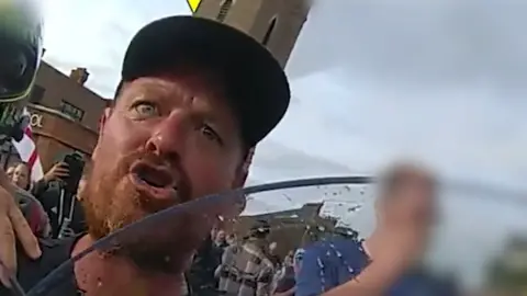 Essex Police A man wearing a black cap alleged to be Lee Gower. He appears to be shouting at a police officer who is behind a riot shield.