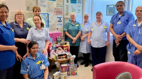 Staff in a hospital are standing around a table which has several toys and games on it. They are all dressed in hospital uniform and smiling. 