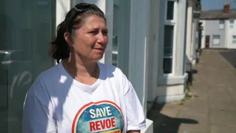 An image of Anna Penfold Sadler outside her house, she is wearing a white t-shirt with save Revoe written on it.  She has brown hair and is white.