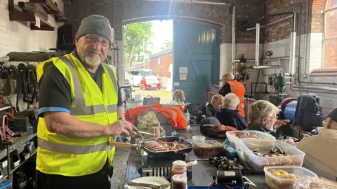 A man in a grey beanie hat, glasses and grey beard is wearing a flourescent vest and appears to be cooking sausages on a frying pan. To the right of the picture a group of men are sat around in wha appears to be a storeroom for tools