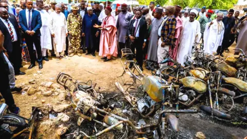 A group of men and officials looking on at the burnt-out remnants of motorbikes 