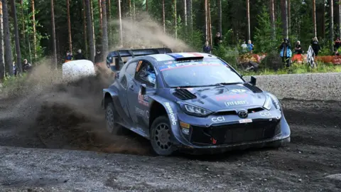 Getty Images A picture of a racing car in a motor sport event. The car is in a forest with brown mud flying up into the sky behind the car. There are people watching the car in the background.