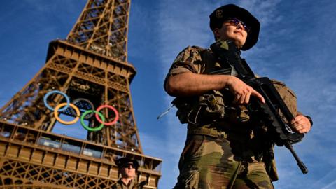 French soldiers walk in Paris holding guns with the Eiffell Tower in the background displaying the Olympic games logo