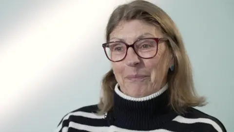 A woman with short light brown hair and glasses smiling to the camera. She is sitting on a chair against a white backdrop and wearing a black and white striped jumper.