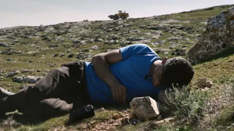 Basel Adra lies on the grass in his home community of Masafer Yatta, his camera beside him, as an Israeli army bulldozer is seen on a rocky hill behind him 