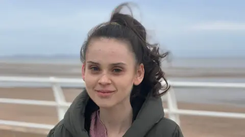 Freya is standing on a coastal path in Rhyl. The beach and sea is behind her. She is looking straight at the camera and has a small smile. She has brown hair that has been tied back because of the windy weather. The sky is very grey and Freya is wearing a large puffer jacket to keep warm. 