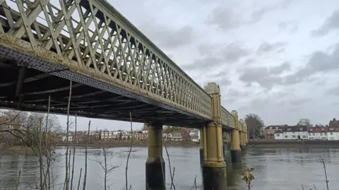 A view of Kew Railway Bridge across the River Thames, with houses on the far river bank and a grey cloudy sky.