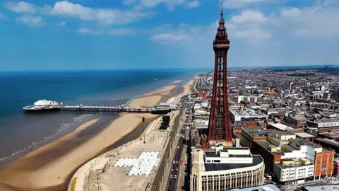 A general aerial view of Blackpool showing the tower and the town to the right, and the beach to the left with central pier. The sky is blue with a few white clouds.