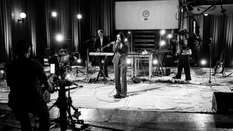 A black and white photo of Charli XCX performing at BBC Maida Vale studios. She is wearing a suit, and there are two people performing behind her. On the left hand side of the photograph, you can see the back of a camera person and a camera filming the performance. 